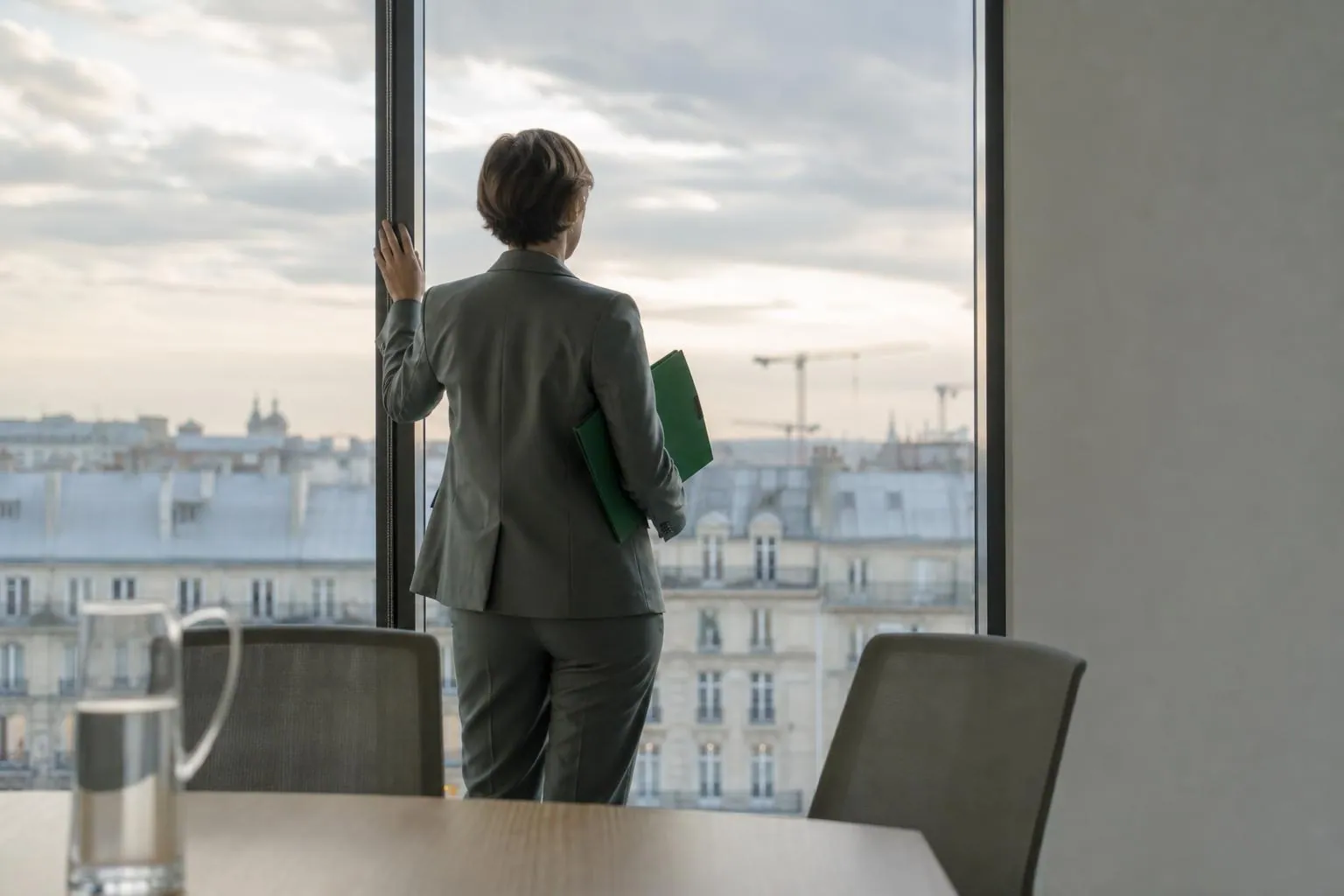 Une femme debout de dos tient un dossier sous le bras devant la baie vitree d un plateau de bureaux donnant sur une ville